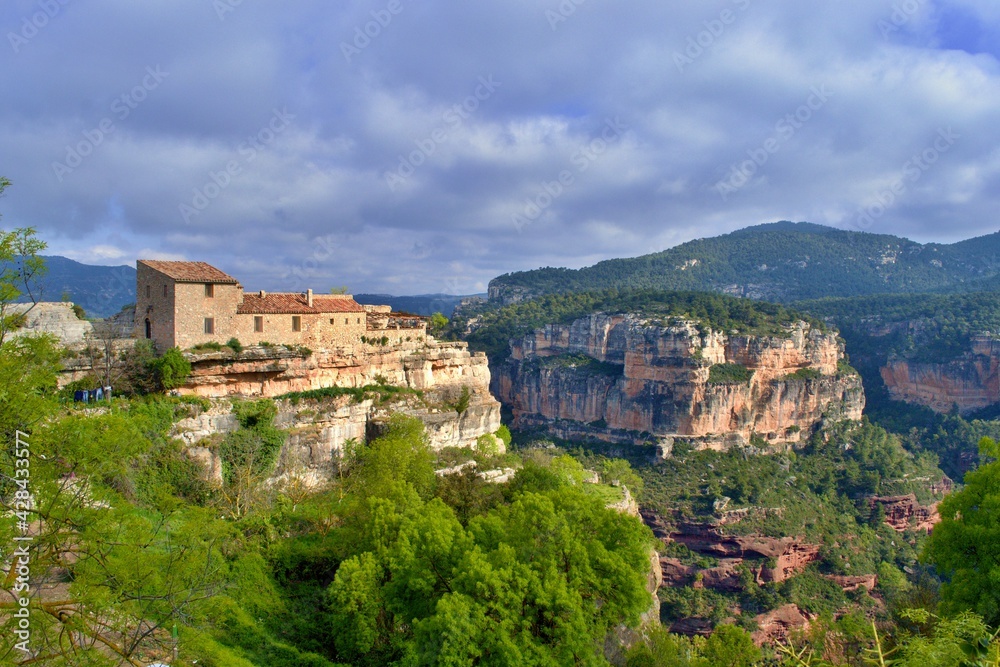 Inn, building, house on the edge of cliff with beautiful view into the valley of red rocks, green bushes. Mountains in the background. Sun, clouds.  Siurana, Catalonia, Spain.