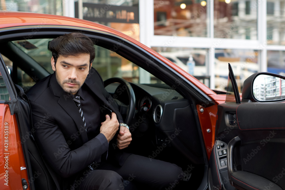 Young caucasian businessman in  black suit step down from a bright sports car while put his mobile phone into suit pocket.
