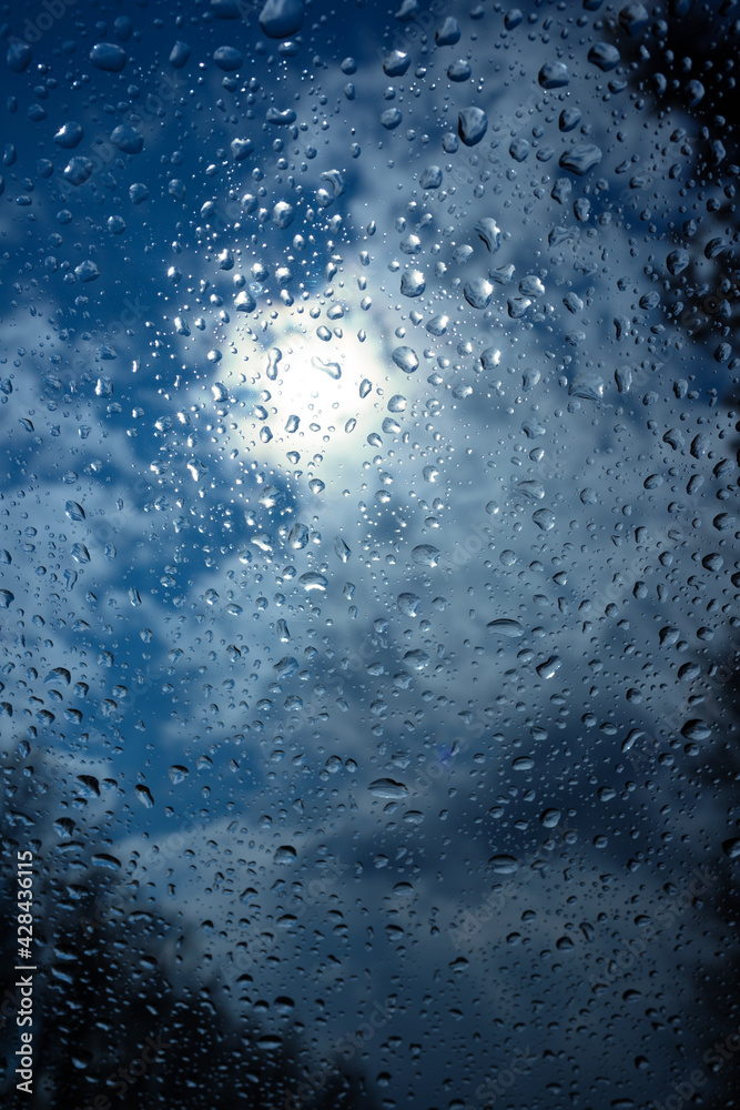 Wet windshield taken from the interior of a car. Rain falls on the surface of a car window glass on a gray background. The sun is shining from behind the clouds. Water drops wallpaper. Wet windshield taken from the interior of a car. Rain falls on the surface of a car window glass on a gray background. The sun is shining from behind the clouds. Water drops wallpaper.