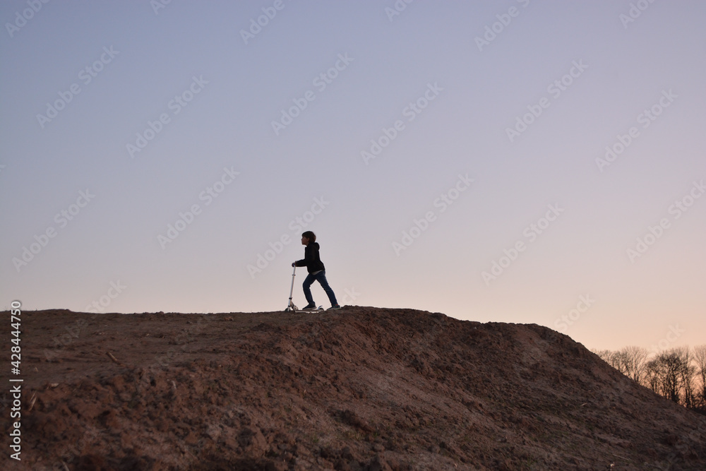 Silhouette of a playing boy with a roller playing with a scooter on a earthy hill at sunset