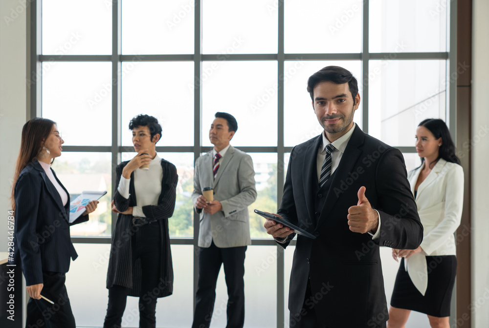 portrait of young caucasian confident businessman with smiling face stnading in office with background of multiethnic colleagues. diversity in business concept portrait of young caucasian confident businessman with smiling face stnading in office with background of multiethnic colleagues. diversity in business concept