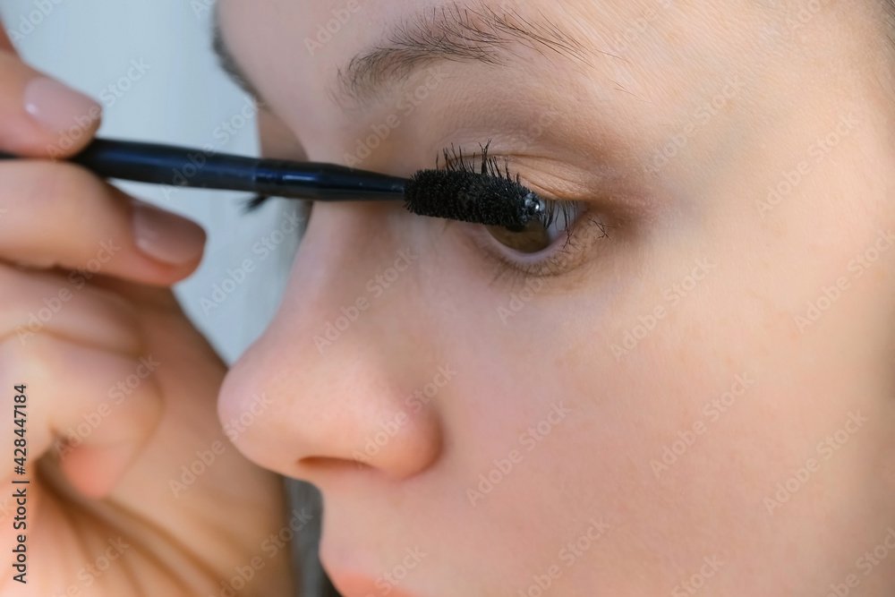 Woman is painting lashes using mascara and brush looking at small mirror, closeup face view. Female is doing makeup. Everyday routine for woman.