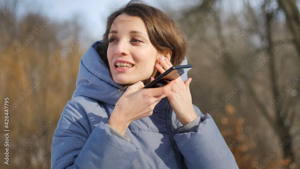 Young woman with wireless headphone is using her smartphone to send a voice message to a new social network Clubhouse standing somewhere outdoors at the park Young woman with wireless headphone is using her smartphone to send a voice message to a new social network Clubhouse standing somewhere outdoors at the park