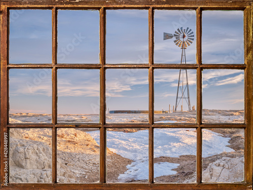 windmill with a pump and cattle water tank in a prairie  winter or early spring scenery as seen from a vintage sash window