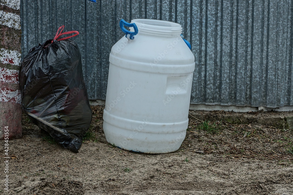 an empty white plastic barrel and a black garbage bag are on the ground near a metal fence wall outside an empty white plastic barrel and a black garbage bag are on the ground near a metal fence wall outside