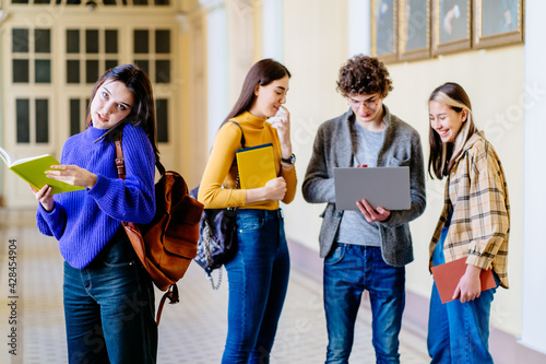 Positive young woman university student with backpack and books talking on cell phone, looking at camera, smiling with friends stand in a university hall during break on background.