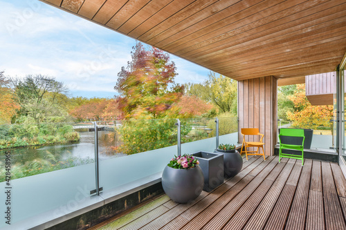 Colorful chairs and pots with flowers placed on terrace near glass fence against river and autumn trees