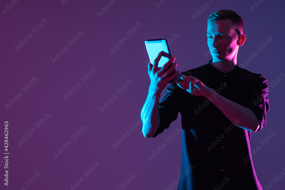 Portrait of a man with a tablet. Student looks at screen of the tablet. Guy is holding gadget in front of him. Student with a tablet on a purple background. Concept - sale of tablets for study. - obrazy, fototapety, plakaty 