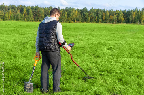Lawnmower in hands of a man. Man with a lawnmower and a shovel in his hands. Human with lawnmower stands with his back to camera. Young guy is mowing grass. Grass processing on a summer day.