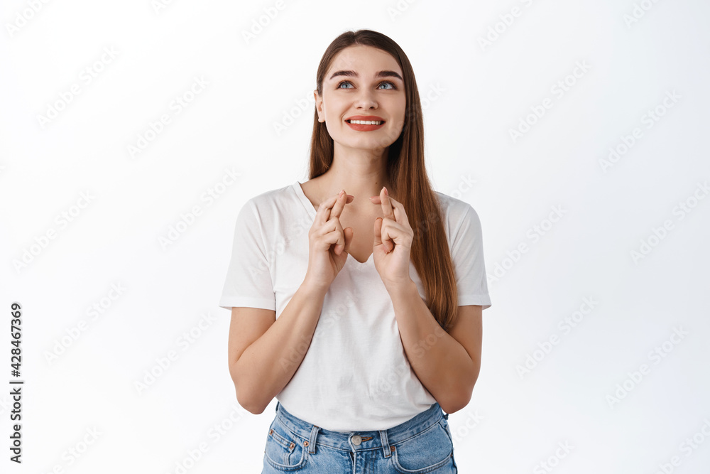 Smiling cute girl making wish, cross fingers for luck and looking up, begging god for good positive results, anticipating news, standing over white background Smiling cute girl making wish, cross fingers for luck and looking up, begging god for good positive results, anticipating news, standing over white background