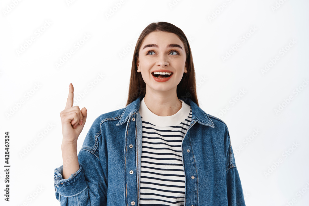 Smiling caucasian female model pointing, looking up with happy pleased face, checking out promotional offer, logo or banner on top copyspace, standing over white background Smiling caucasian female model pointing, looking up with happy pleased face, checking out promotional offer, logo or banner on top copyspace, standing over white background
