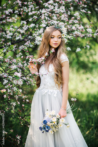 Attractive young woman in white wedding dress with bouquet of wild flowers walks in the summer garden, enjoying the blooming spring nature