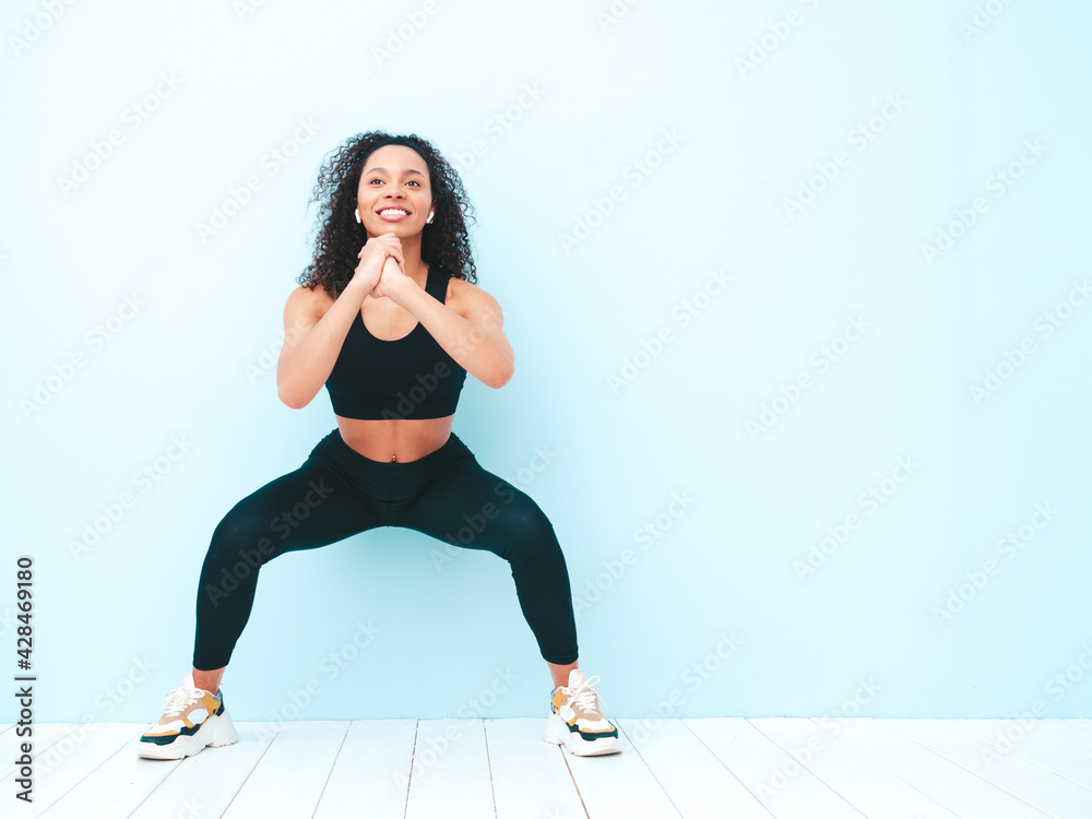Fitness smiling black woman in sports clothing with afro curls hairstyle.She doing squats. Young beautiful model with perfect tanned body.Female in studio near light blue wall Fitness smiling black woman in sports clothing with afro curls hairstyle.She doing squats. Young beautiful model with perfect tanned body.Female in studio near light blue wall
