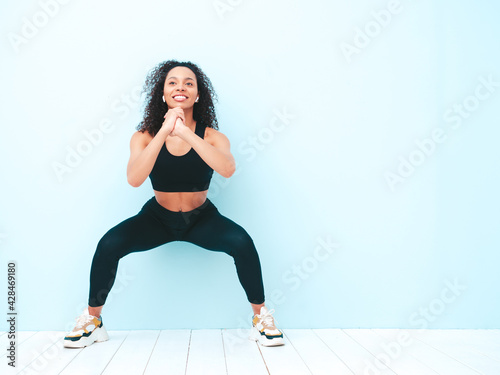 Fitness smiling black woman in sports clothing with afro curls hairstyle.She doing squats. Young beautiful model with perfect tanned body.Female in studio near light blue wall