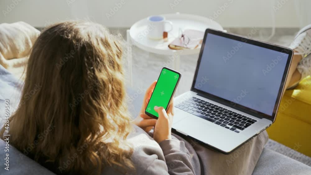 Brunette woman looks at phone with chromakey screen holding contemporary laptop on knees on sofa in living room in sunny morning backside view