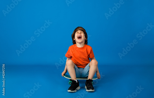 Canvas Print cheerful little boy with brown hair in a T-shirt and denim shorts sits on a skateboard on a blue background with space for text