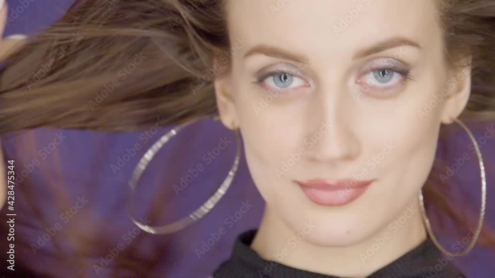 Charming beautiful cute young girl brunette model with beautiful eyes poses for camera and touches wind blowing hair on purple background in foto studio Charming beautiful cute young girl brunette model with beautiful eyes poses for camera and touches wind blowing hair on purple background in foto studio