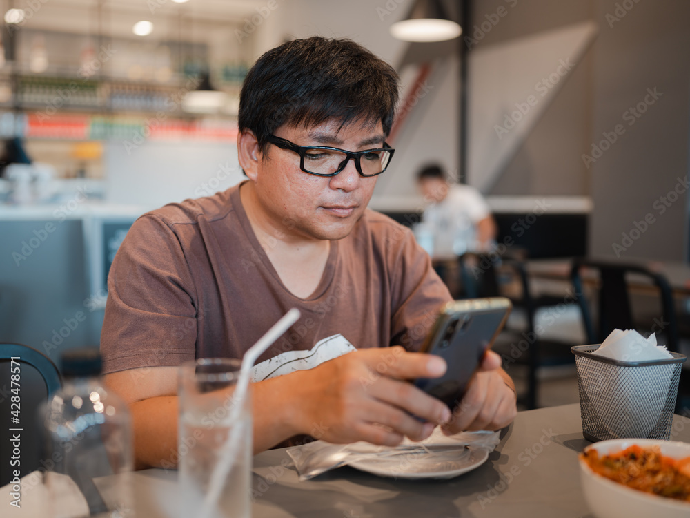 Asian man with eyeglasses watching at smartphone during eating in restaurant, mobile phone addicted, working outside office 