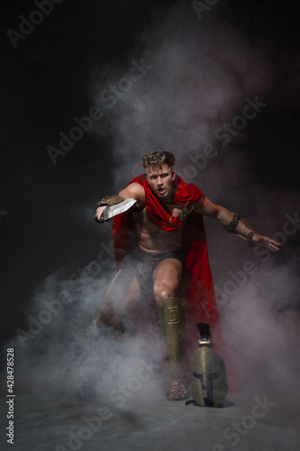 Young athletic sexy man in a costume of a Roman warrior in a red cloak in move on a black background in the smoke.