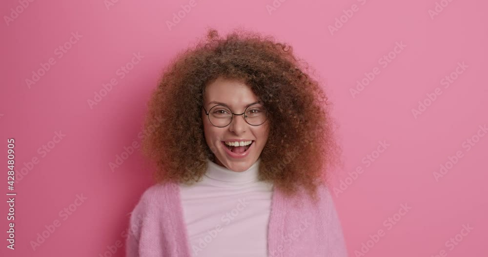 Beautiful cheerful woman with curly bushy hair points directly at camera demonstrates something funny wears round spectacles casual sweater isolated over pink background notices comic scene. Beautiful cheerful woman with curly bushy hair points directly at camera demonstrates something funny wears round spectacles casual sweater isolated over pink background notices comic scene.