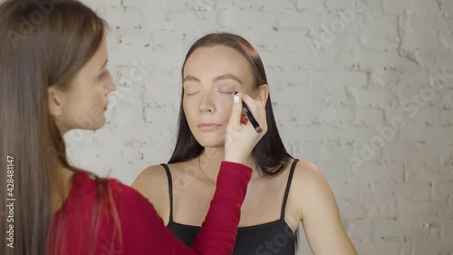 Professional makeup artist girl makes beautiful makeup for charming young model girl with charming eyes in studio against of white brick wall