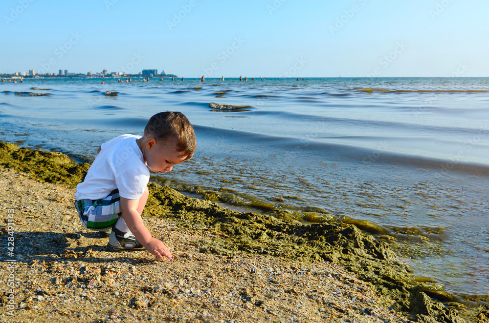 Emotional portrait of a child brightly lit. Looks concentrated, squints from the sun. Wearing a hat against the blue sea and sky, on the shore on a hot summer day.