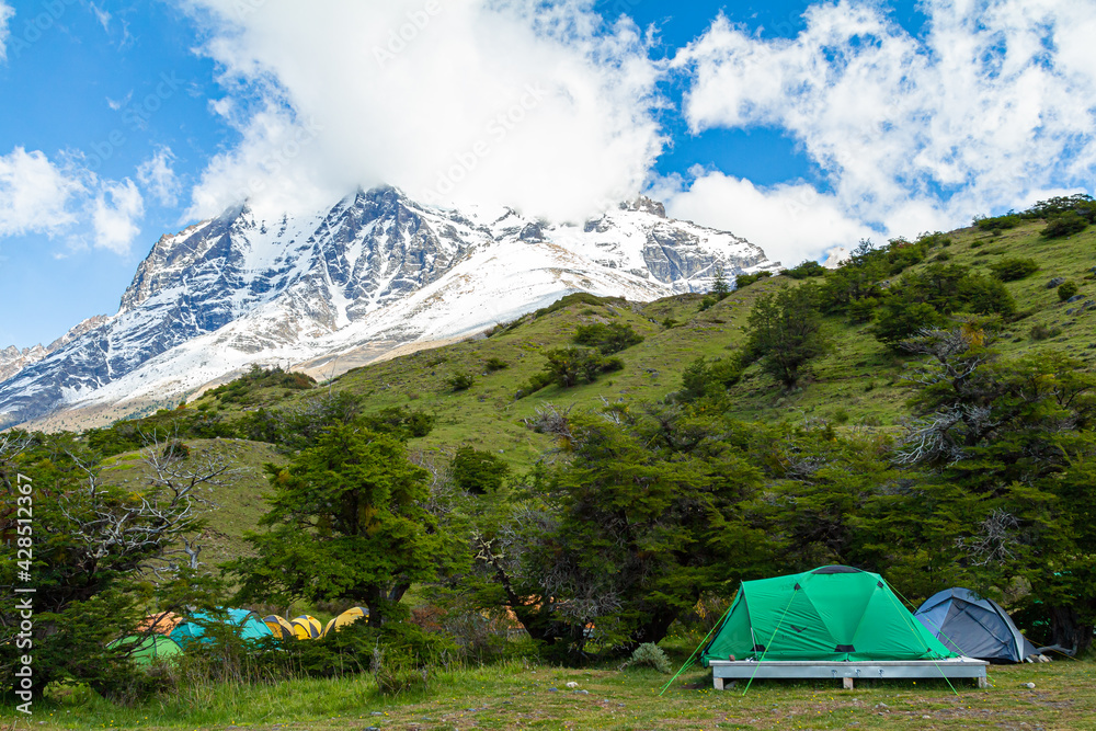 Camping em meio a vegetação nativa com duas barracas verdes e azuis em primeiro plano, montanhas nevadas ao fundo e céu muito azul com nuvens
