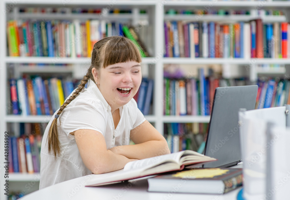 Happy young girl with syndrome down uses a laptop at library. Education for disabled children concept Happy young girl with syndrome down uses a laptop at library. Education for disabled children concept