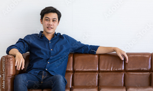 Young man in blue shirt and blue jeans is sitting on a dark brown sofa with smile. He stretch his left arm out for relax.