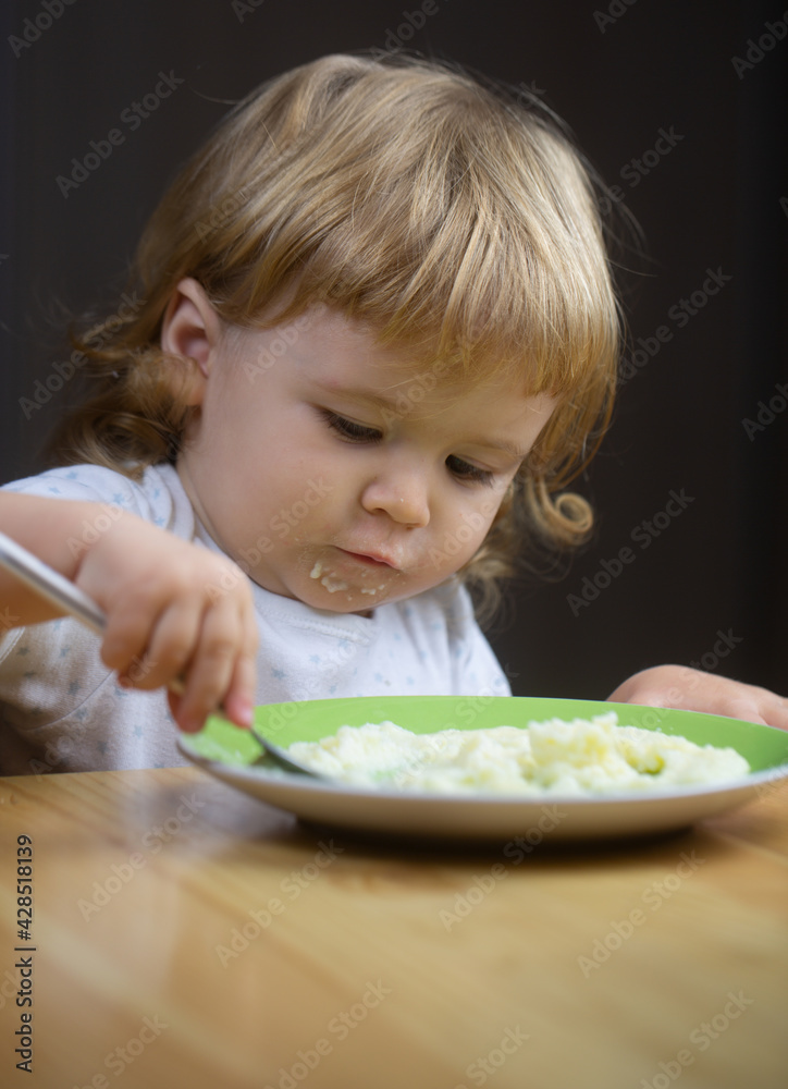 Child eating, nutrition concept. Cute baby face with a spoon and a plate in the kitchen at home. Child eating, nutrition concept. Cute baby face with a spoon and a plate in the kitchen at home.
