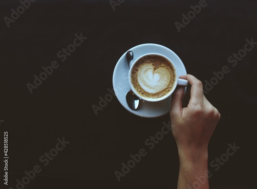 White cup of espresso coffee with a womans hand holding the handle. Small spoon and saucer. Black background with copyspace for text. Dark concept. Strong coffee
