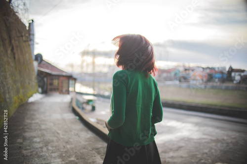 Back view of a skinny redhead girl wearing a green jacket and a wind blowing her hair