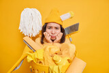 Displeased sad woman doesnt want to clean room looks sadly at mess and dirt uses different cleaning tools poses near laundry basket against yellow background dressed casually. Dejected housemaid