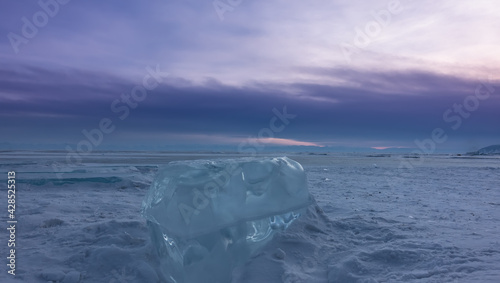 Shiny turquoise transparent ice floe on the snow-covered surface of a frozen lake. The background is the sunset lilac sky. Baikal