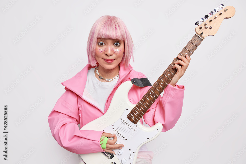 Fototapeta premium Indoor shot of popular female artist plays rock music on electric guitar poses against white background with musical instrument wears pink jacket has glitters on face. Popular singer has rehearsal