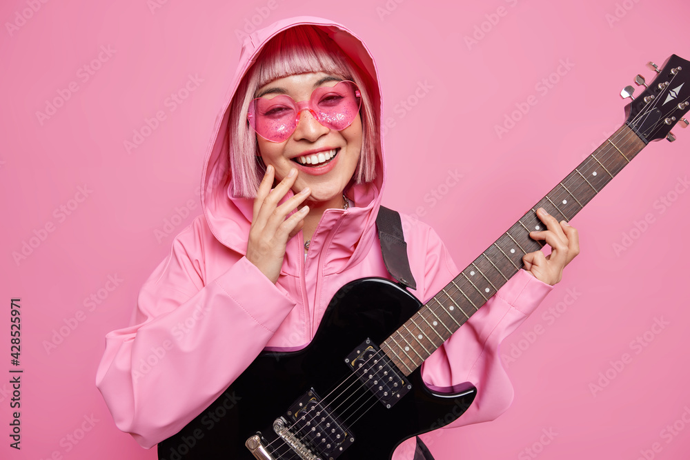 Fototapeta premium Pleased fashionable woman dressed in stylish jacket pink sunglasses has fun pretends to be rock star smiles broadly uses electric guitar isolated over rosy background. Talented musician indoor