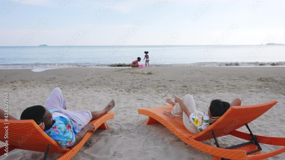 Happy mixed race family holiday vacation. Parents resting on beach chair looking at two cute child girl playing sand together on the beach in summer day. Family relax and having fun in summer weekend Happy mixed race family holiday vacation. Parents resting on beach chair looking at two cute child girl playing sand together on the beach in summer day. Family relax and having fun in summer weekend
