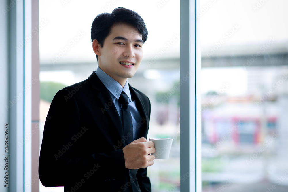 Asian business man wearing blue shirt, formal suit with necktie, happily smiling with brace on his teeth and holding a white cup of coffee while standing beside the window in office in the morning. Asian business man wearing blue shirt, formal suit with necktie, happily smiling with brace on his teeth and holding a white cup of coffee while standing beside the window in office in the morning.