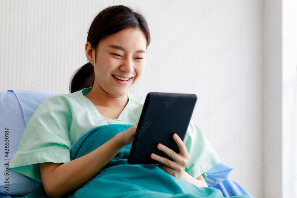 An asian young pretty female patient smiling with happiness and touching screen of tablet for online chat or communicate while lying on bed in hospital. Technology and Medical Concept. An asian young pretty female patient smiling with happiness and touching screen of tablet for online chat or communicate while lying on bed in hospital. Technology and Medical Concept.