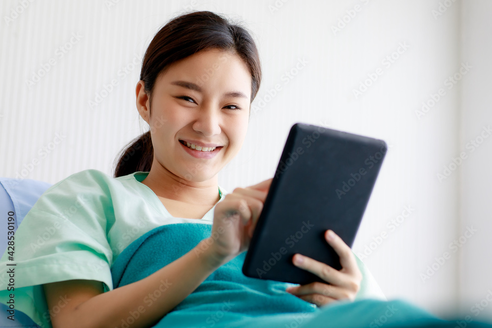 An asian young pretty female patient smiling with happiness and touching screen of tablet for online chat or communicate while lying on bed in hospital. Technology and Medical Concept. An asian young pretty female patient smiling with happiness and touching screen of tablet for online chat or communicate while lying on bed in hospital. Technology and Medical Concept.
