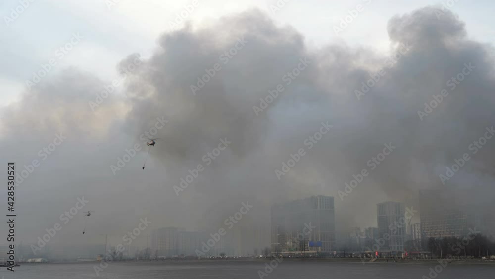 Helicopters fly with bambi bucket. Rescue helicopters over river. City is in smoke. Fire. Houses on embankment in smoke
