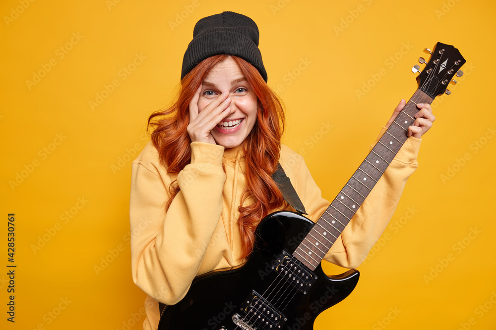 Fototapeta premium Happy female guitar player with natural red hair chuckles and keeps hand on face plays favorite genre of music holds black electric guitar dressed in stylish clothes isolated over yellow background