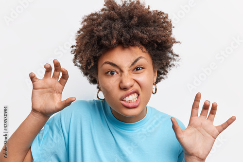 Headshot of Afro American woman makes paws gesture roars or growls like animal pretends being cat clenches teeth wears casual blue t shirt isolated over white background. Facial expressions.