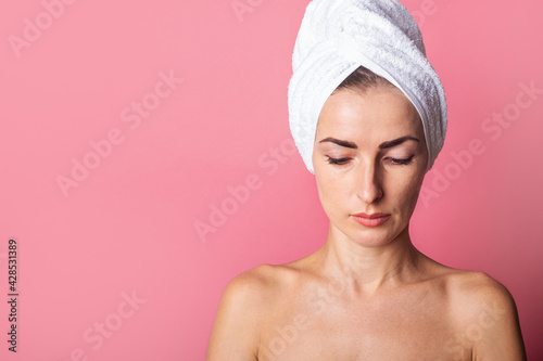 young woman with a towel on her head, nude shoulders, looks down on a pink background