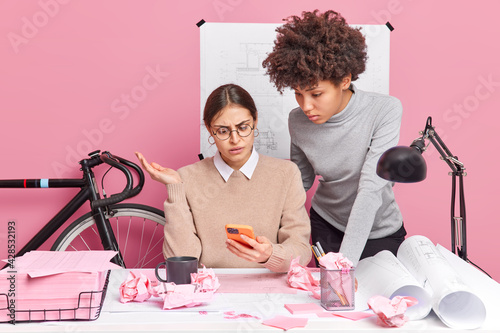 Two diverse women wotk at office desk on new promotion action use mobile phone look with indignation at display cannot understand what mistake they did have mess at dekstop. Freelancers working