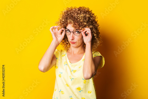 curly young woman in a T-shirt looks from under glasses at the camera on a yellow background