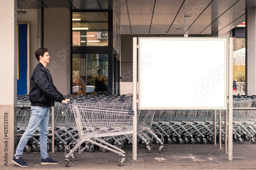Young person driving the empty shopping cart to a supermarket in front of blank billboard.