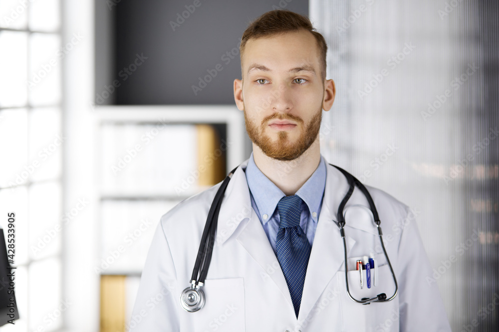 Red-bearded doctor standing straight in clinic near his working place. Portrait of physician. Medicine, healthcare Red-bearded doctor standing straight in clinic near his working place. Portrait of physician. Medicine, healthcare