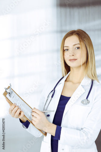 Young intelligent woman-doctor is prescribing some medicine for a patient, using a clipboard, while standing in the cabinet in a clinic. Female physician with a stethoscope at her working place