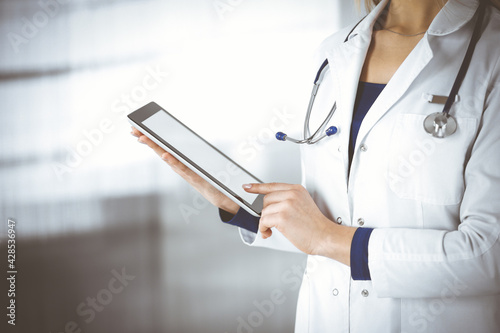 Unknown woman-doctor is holding a tablet computer in her hands, while standing in a clinic. Female physician with a stethoscope in her office, close-up. Perfect medical service in a hospital. Medicine